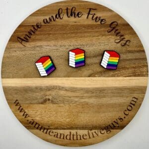 A wooden circular board with "Annie and the Five Guys" written on it, featuring three rainbow-colored pin buttons shaped like stacks of books. The website "www.annieandthefiveguys.com" is at the bottom.