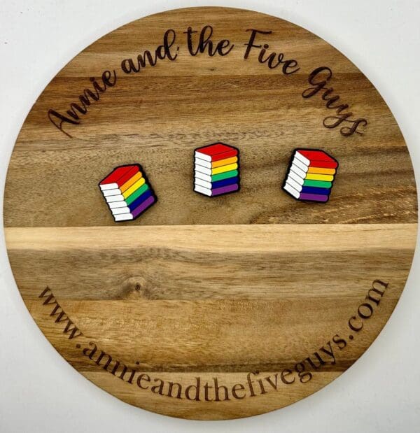 A wooden circular board with "Annie and the Five Guys" written on it, featuring three rainbow-colored pin buttons shaped like stacks of books. The website "www.annieandthefiveguys.com" is at the bottom.