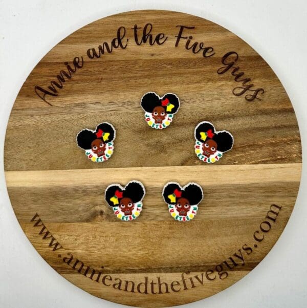 A round wooden board with "Annie and the Five Guys" engraved around the edge displays five pins featuring a cartoon character with large hair, a red flower, and multicolored clothing.