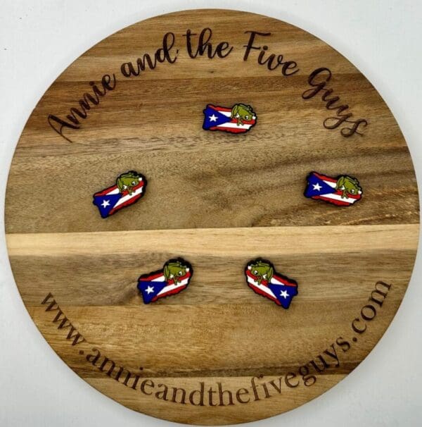 A wooden round sign reading "Annie and the Five Guys" with the website "www.annieandthefiveguys.com." It displays five small Puerto Rican Flag silicone focal beads featuring a green character over a flag design.