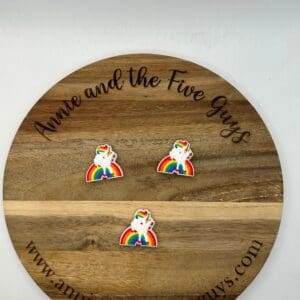 A round wooden plaque with "Annie and the Five Guys" and website text around the edge. Three rainbow unicorn pins are displayed on the surface.