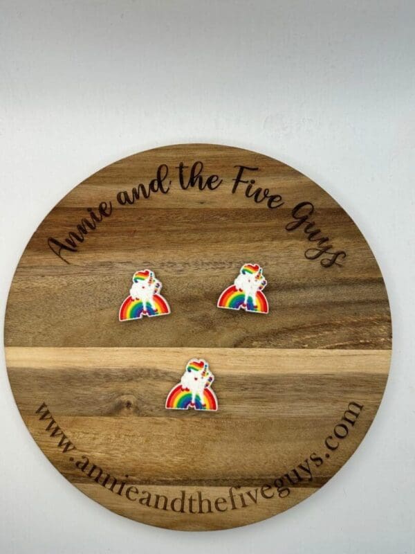 A round wooden plaque with "Annie and the Five Guys" and website text around the edge. Three rainbow unicorn pins are displayed on the surface.