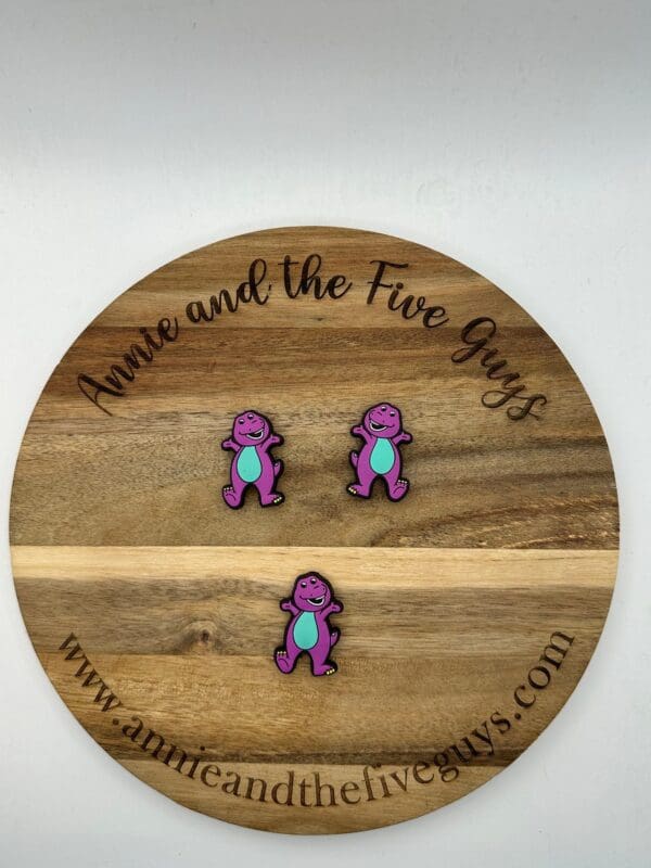 Wooden round with "Annie and the Five Guys" engraved, showcasing three purple and turquoise dinosaur pins. Website "www.annieandthefiveguys.com" is visible at the bottom.