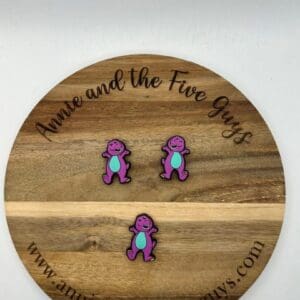 Wooden round with "Annie and the Five Guys" engraved, showcasing three purple and turquoise dinosaur pins. Website "www.annieandthefiveguys.com" is visible at the bottom.