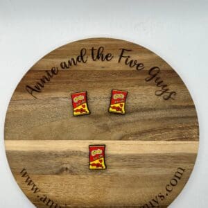 Circular wooden board with "Buckeys silicone focal bead (Copy)" engraved, holding three small pins resembling packets of Chili's chips. Website URL "www.annieandthefiveguys.com" is at the bottom of the board.