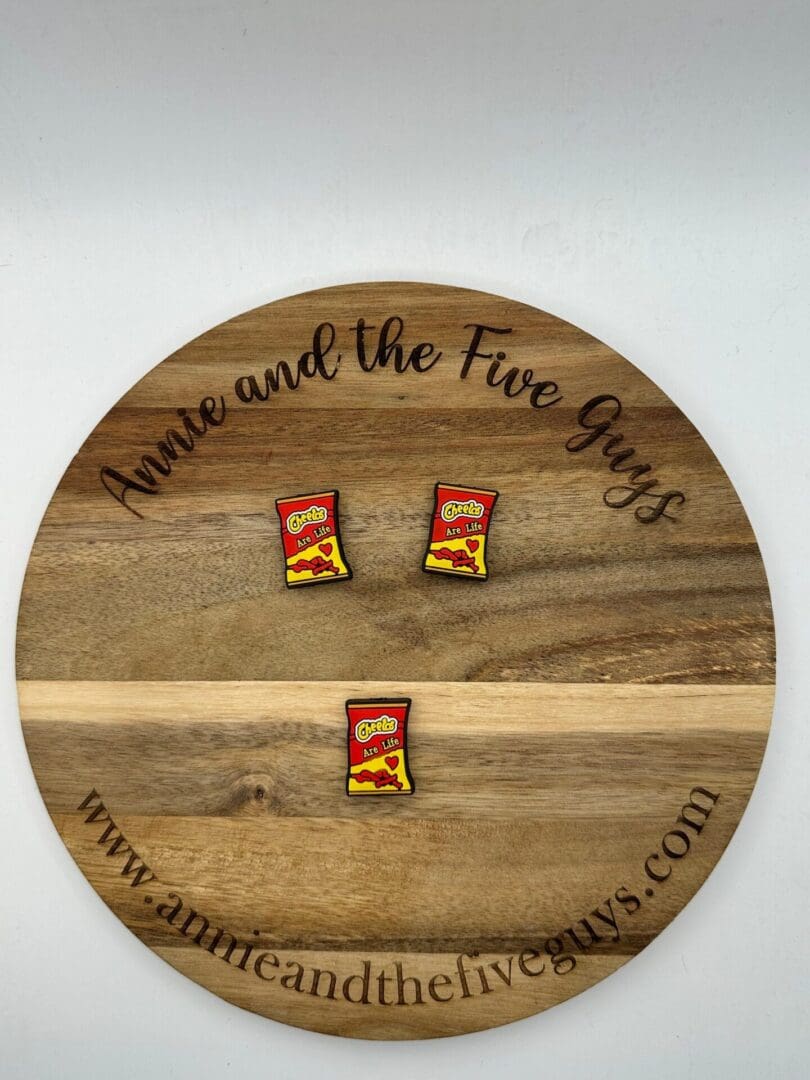 Circular wooden board with "Buckeys silicone focal bead (Copy)" engraved, holding three small pins resembling packets of Chili's chips. Website URL "www.annieandthefiveguys.com" is at the bottom of the board.