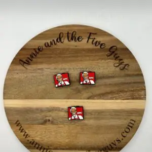 A wooden circular board displaying two KFC logo buttons. The board has the text "Annie and the Five Guys" and the website "www.annieandthefiveguys.com.