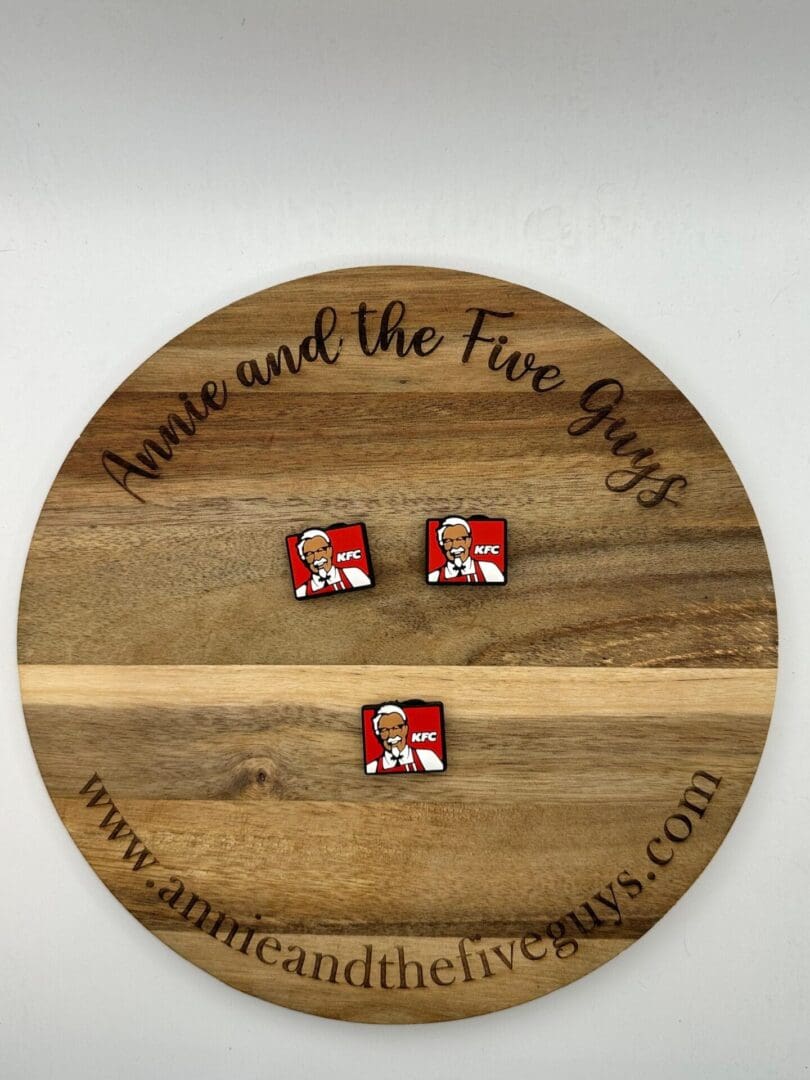 A wooden circular board displaying two KFC logo buttons. The board has the text "Annie and the Five Guys" and the website "www.annieandthefiveguys.com.