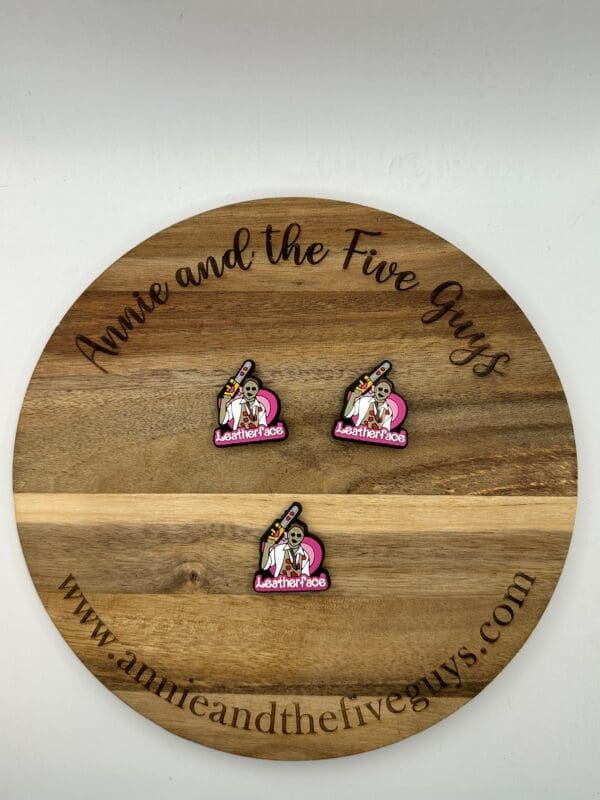 A round wooden board with "Annie and the Five Guys" engraved on it. Three small pins with a cartoon character and the word "Leather face silicone focal bead" are displayed on the board.