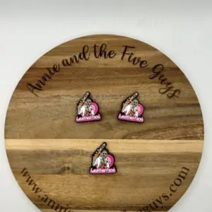 A round wooden board with "Annie and the Five Guys" engraved on it. Three small pins with a cartoon character and the word "Leather face silicone focal bead" are displayed on the board.