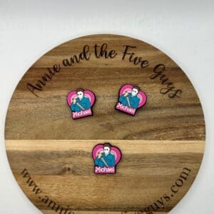 A round wooden board with "Annie and the Five Guys" text and three identical pink pins showing a cartoon figure named "Michael" in front of a heart background.