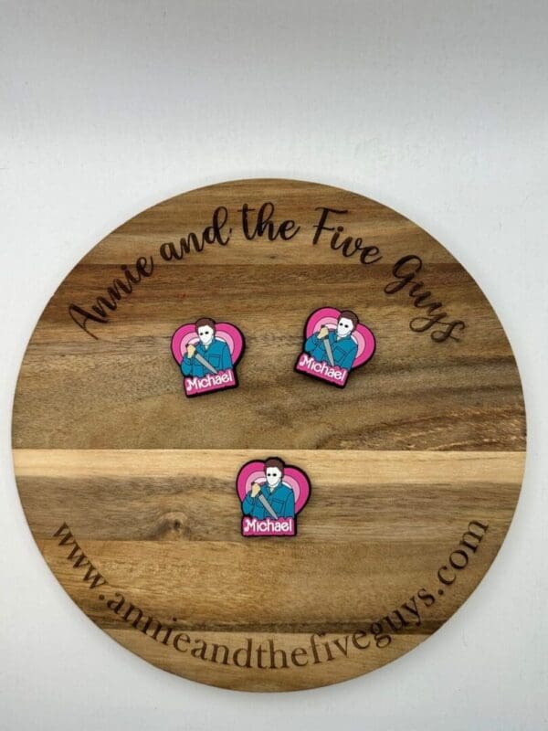 A round wooden board with "Annie and the Five Guys" text and three identical pink pins showing a cartoon figure named "Michael" in front of a heart background.