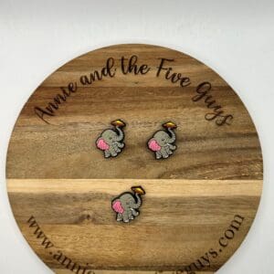 Two elephant-themed pins with yellow hats on a round wooden surface displaying the text "Annie and the Five Guys" and the website "www.annieandthefiveguys.com.