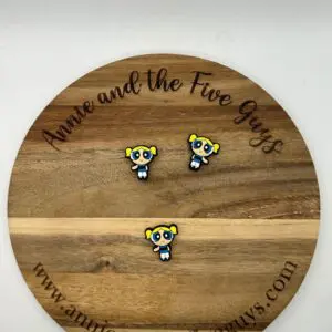 A round wooden plaque with "Annie and the Five Guys" written on the top and bottom, displaying three identical pins of a cartoon character with yellow pigtails, large blue eyes, and a white dress.