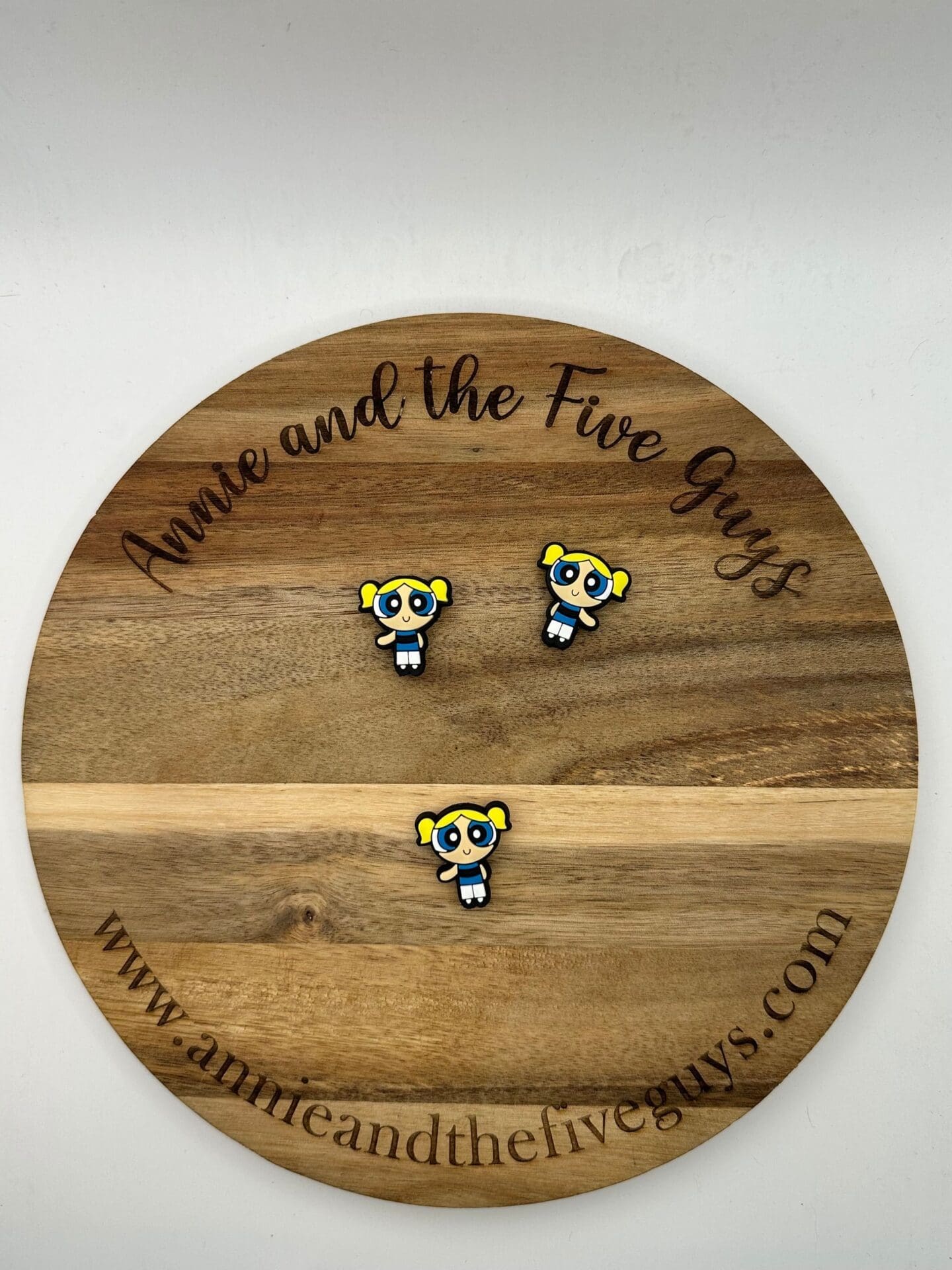 A round wooden plaque with "Annie and the Five Guys" written on the top and bottom, displaying three identical pins of a cartoon character with yellow pigtails, large blue eyes, and a white dress.