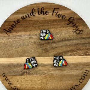 A wooden round surface with three pins depicting a bearded figure in a robe holding a rainbow-colored garment. The text on the pins reads "Y'all Need Me?" The surface is marked with "Annie and the Five Guys" and a website.