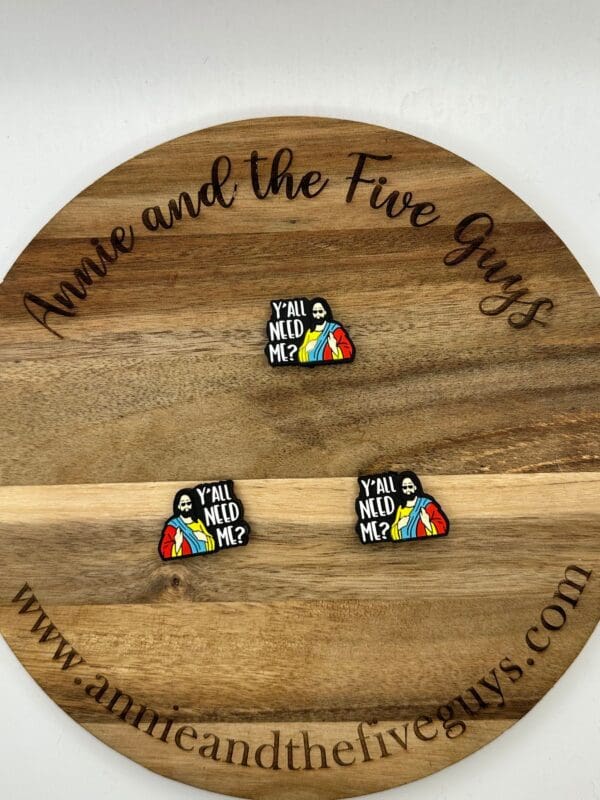 A wooden round surface with three pins depicting a bearded figure in a robe holding a rainbow-colored garment. The text on the pins reads "Y'all Need Me?" The surface is marked with "Annie and the Five Guys" and a website.