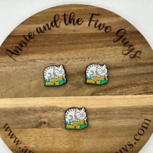 Three enamel pins featuring a cat sitting on books placed on a wooden circular board with the text "Annie and the Five Guys" and "www.annieandthefiveguys.com" engraved around the edge.