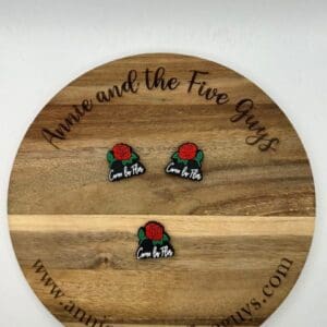 A round wooden board with black text that reads "Annie and the Five Guys" in a circular pattern and "www.annieandthefiveguys.com" at the bottom. Three small pins with roses and text "Came by Plan" are on the board.