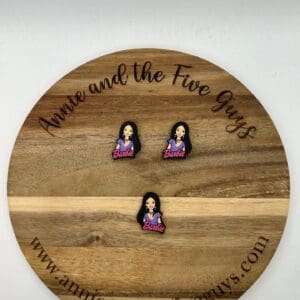 A round wooden board with "Annie and the Five Guys" text holds three identical pins featuring a figure with long black hair and a shirt labeled "Barbie." Website URL shown at the bottom.