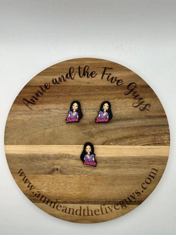 A round wooden board with "Annie and the Five Guys" text holds three identical pins featuring a figure with long black hair and a shirt labeled "Barbie." Website URL shown at the bottom.