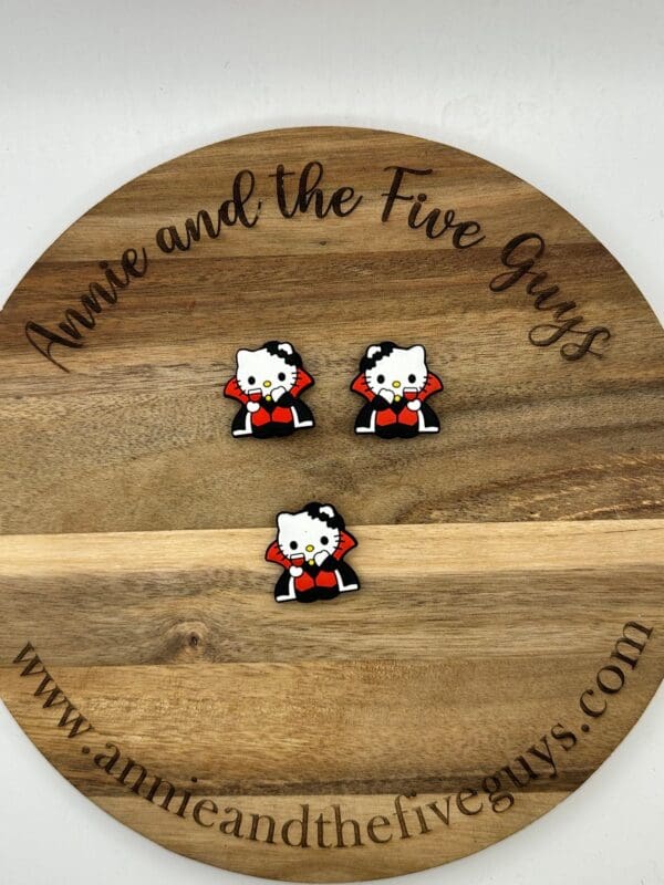 A round wooden board with the text "Annie and the Five Guys" holds three small pins featuring a cat in a red cape. The bottom of the board includes the website "www.annieandthefiveguys.com".