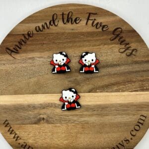 A round wooden board with the text "Annie and the Five Guys" holds three small pins featuring a cat in a red cape. The bottom of the board includes the website "www.annieandthefiveguys.com".