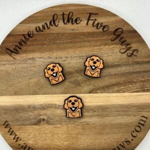 Three dog-shaped pins are displayed on a round wooden board with engraved text reading "Annie and the Five Guys" and the website "www.annieandthefiveguys.com".