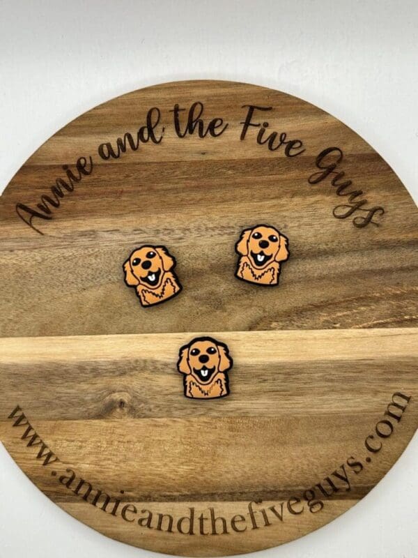 Three dog-shaped pins are displayed on a round wooden board with engraved text reading "Annie and the Five Guys" and the website "www.annieandthefiveguys.com".