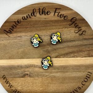 Three small enamel pins featuring a character with blonde hair, sunglasses, and a teal shirt arranged on a wooden circular plaque that reads "Annie and the Five Guys - www.annieandthefiveguys.com".