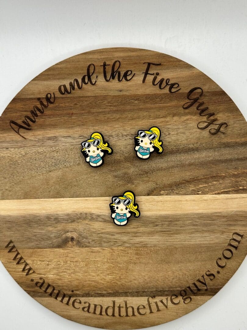 Three small enamel pins featuring a character with blonde hair, sunglasses, and a teal shirt arranged on a wooden circular plaque that reads "Annie and the Five Guys - www.annieandthefiveguys.com".