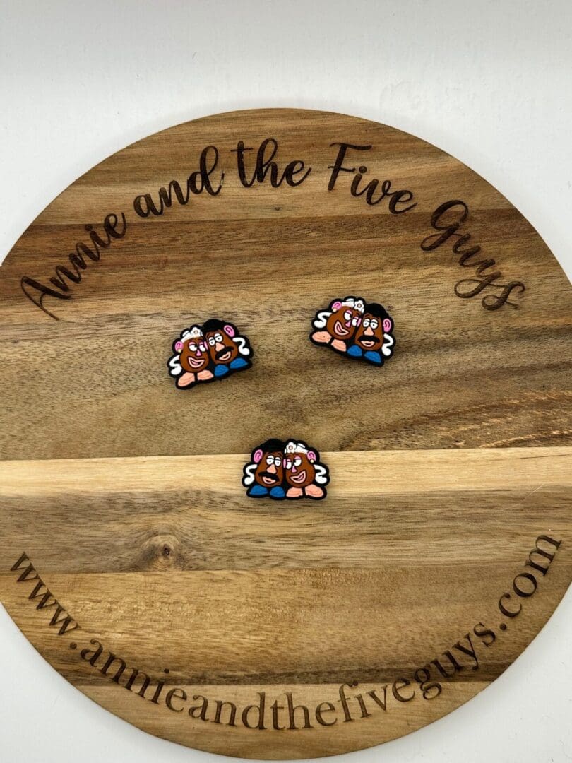 A wooden board engraved with "Annie and the Five Guys" featuring three small, cartoon-like pins resembling Mr. And Mrs. Potatoes Head silicone focal beads.