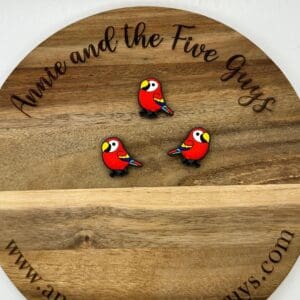 Three small red parrot pins are displayed on a round wooden board engraved with "Annie and the Five Guys" and the website "www.annieandthefiveguys.com".