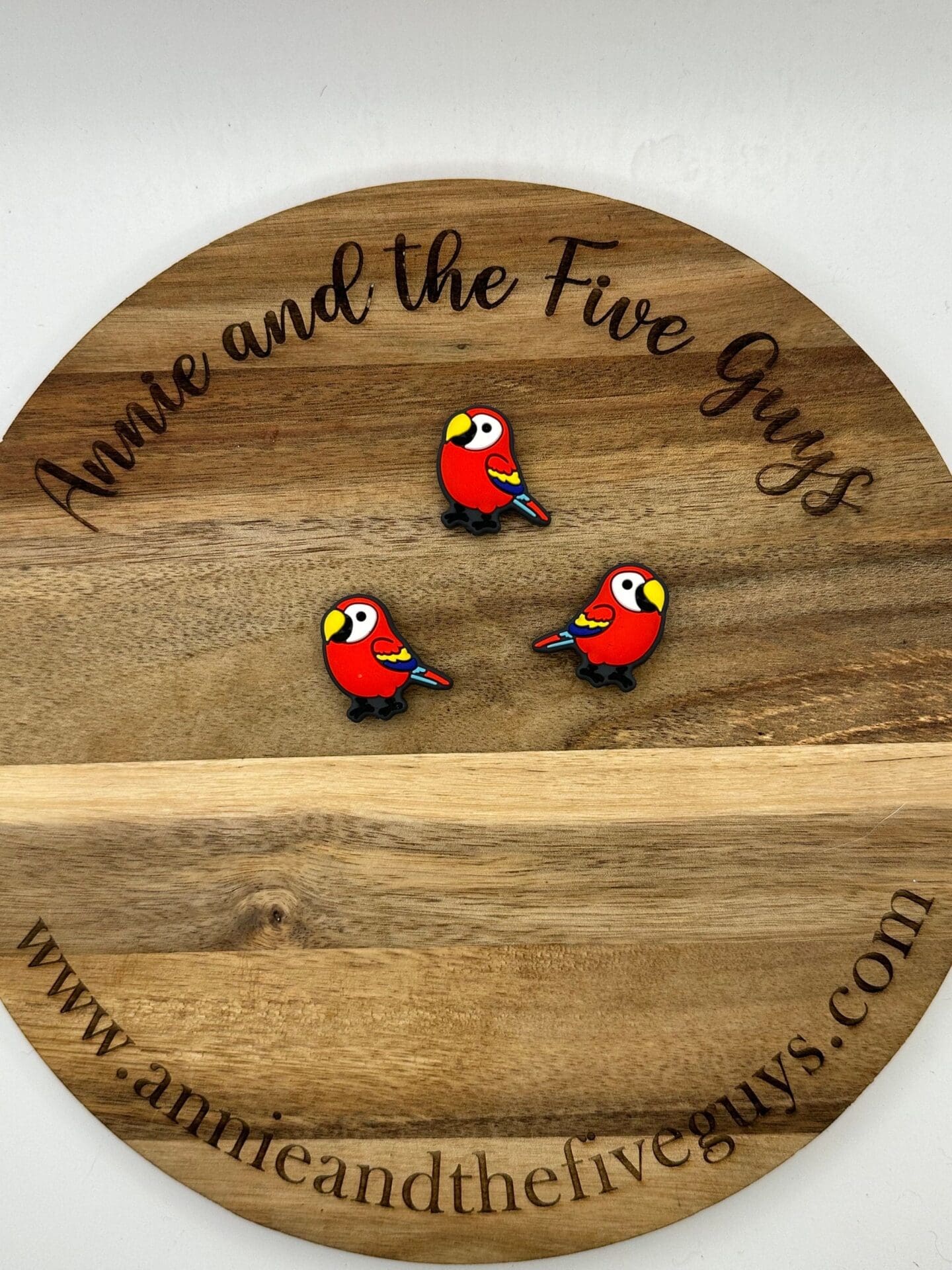 Three small red parrot pins are displayed on a round wooden board engraved with "Annie and the Five Guys" and the website "www.annieandthefiveguys.com".