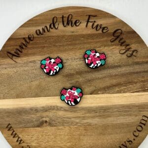 Three pins with the word "Mama" in pink script, floral and cow print designs, are displayed on a wooden board that reads "Annie and the Five Guys" around the edge. Website: www.annieandthefiveguys.com.