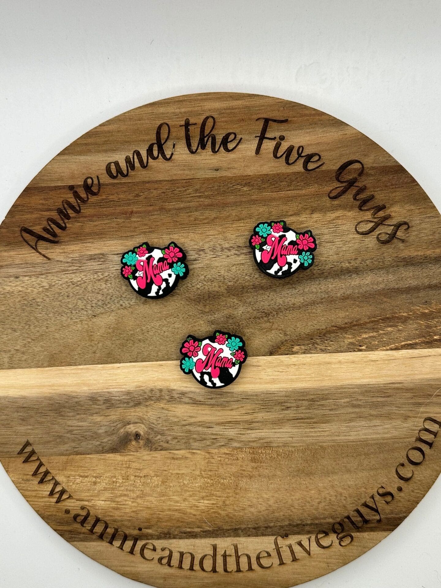 Three pins with the word "Mama" in pink script, floral and cow print designs, are displayed on a wooden board that reads "Annie and the Five Guys" around the edge. Website: www.annieandthefiveguys.com.