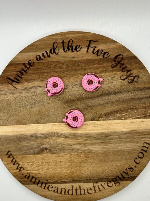 Three pink donut-shaped objects on a wooden board with the text "Annie and the Five Guys" and the website "www.annieandthefiveguys.com".