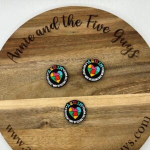 A wooden board with the text "Annie and the Five Guys" and three identical circular pins reading "Autism: A different way of seeing the world." The website "www.annieandthefiveguys.com" is displayed.