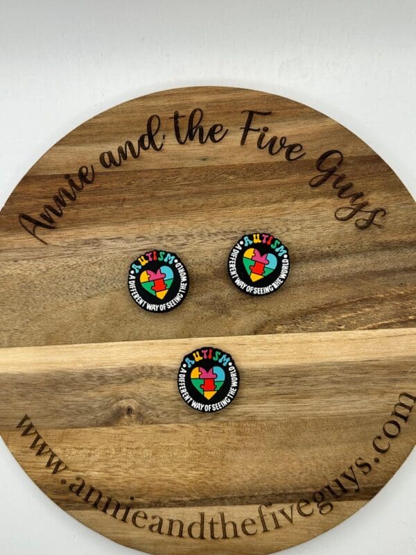 A wooden board with the text "Annie and the Five Guys" and three identical circular pins reading "Autism: A different way of seeing the world." The website "www.annieandthefiveguys.com" is displayed.