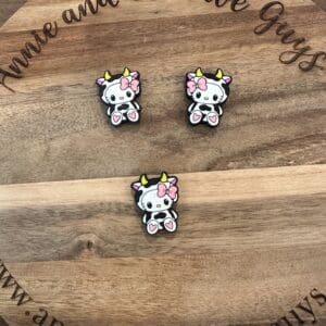 Three cow-shaped pins with pink bows and heart designs on a wooden surface with "Annie and the Five Guys" written around the edge.