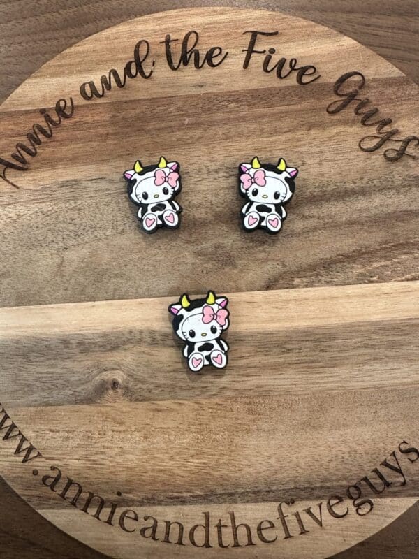 Three cow-shaped pins with pink bows and heart designs on a wooden surface with "Annie and the Five Guys" written around the edge.