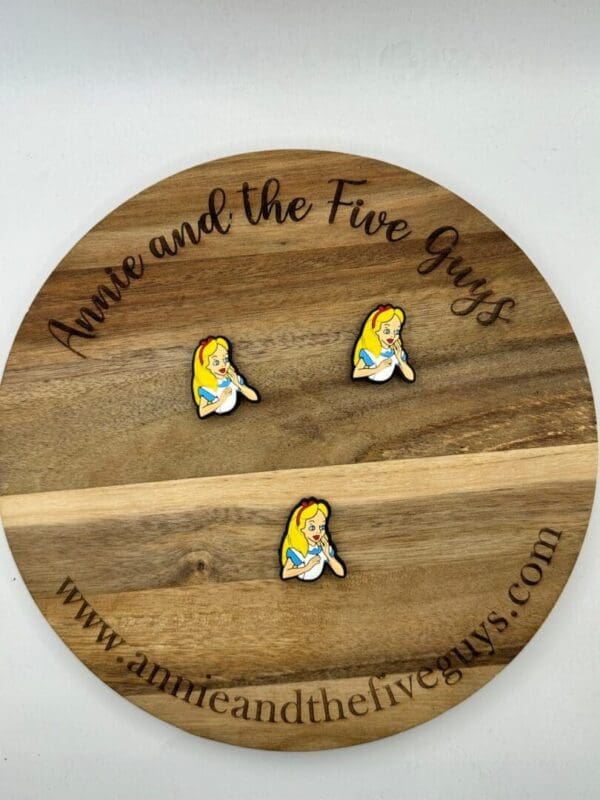 A circular wooden display with "Annie and the Five Guys" text holds three pins depicting a cartoon character with blonde hair and a blue dress. Website www.annieandthefiveguys.com is at the bottom.