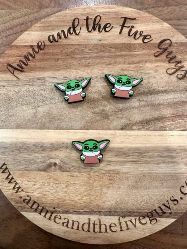 Three pins featuring a green character with large ears and wearing a pink shirt placed on a wooden circular board engraved with "Annie and the Five Guys" and a corresponding website URL.