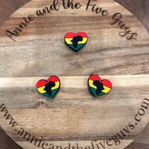 Wooden board with text "Annie and the Five Guys" and website "www.annieandthefiveguys.com" with three heart-shaped pins featuring a black Africa silhouette over red, yellow, and green stripes.