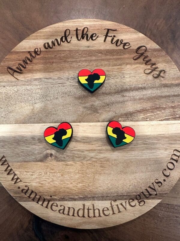 Wooden board with text "Annie and the Five Guys" and website "www.annieandthefiveguys.com" with three heart-shaped pins featuring a black Africa silhouette over red, yellow, and green stripes.