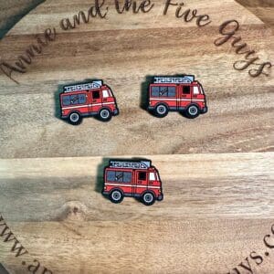 Three fire truck-shaped pins lie on a wooden circular board. The board has cursive text reading "annie and the Five Guys" around its edge.