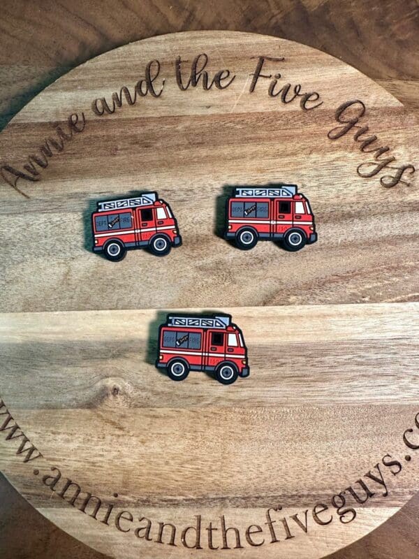 Three fire truck-shaped pins lie on a wooden circular board. The board has cursive text reading "annie and the Five Guys" around its edge.