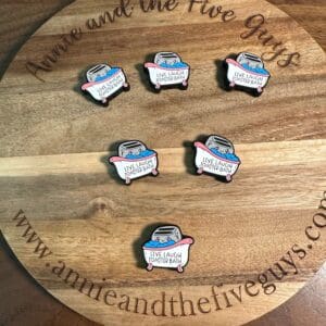 Six enamel pins with the text "Live Laugh Roasted Beans" displayed on a wooden board with "www.annieandthefiveguys.com" and "Annie and the Five Guys" written around the edges.