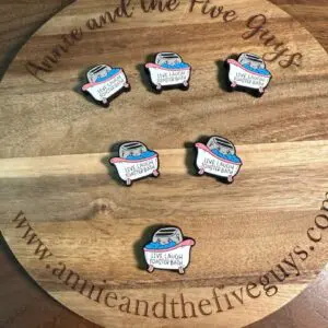 Six enamel pins with the text "Live Laugh Roasted Beans" displayed on a wooden board with "www.annieandthefiveguys.com" and "Annie and the Five Guys" written around the edges.