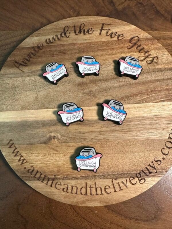 Six enamel pins with the text "Live Laugh Roasted Beans" displayed on a wooden board with "www.annieandthefiveguys.com" and "Annie and the Five Guys" written around the edges.